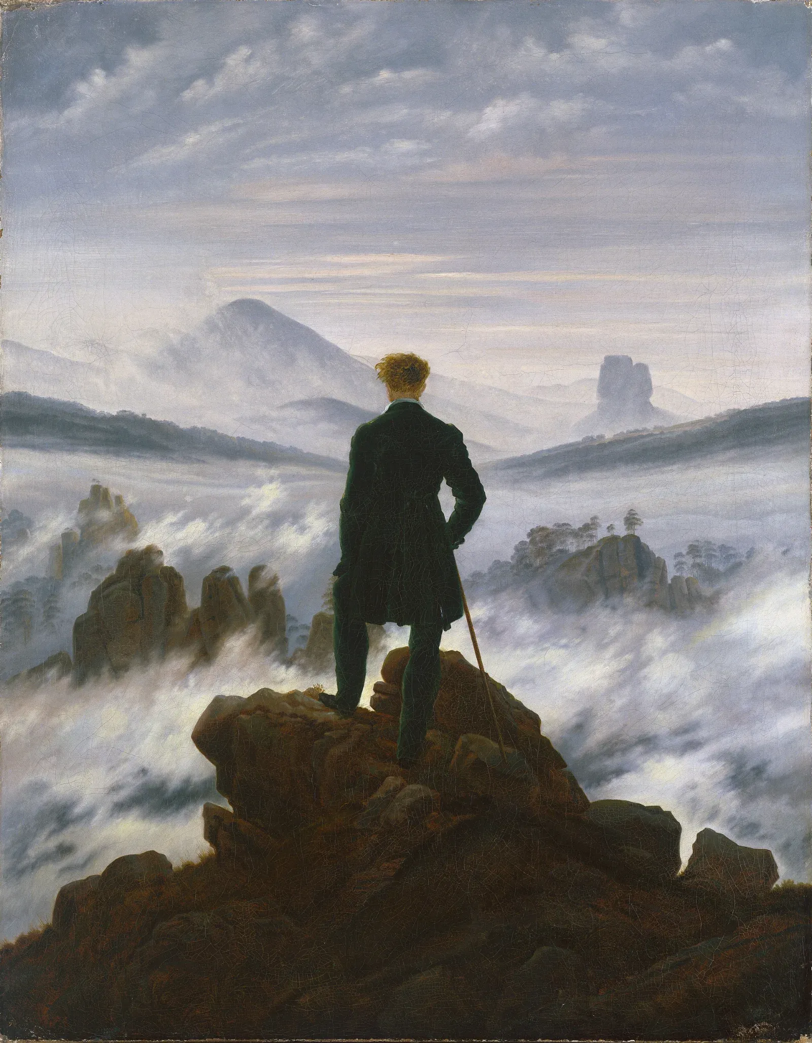 A man in a dark green coat stands with his back to us on a rocky peak, looking out over a landscape of fog, distant rocks and mountains.